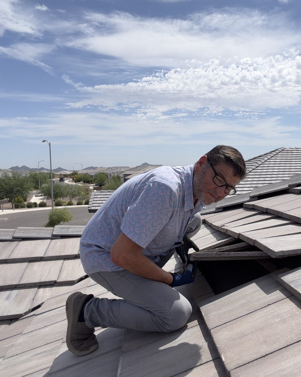 Home inspector Kelly Lopez of Leading Edge Plus Inspections kneeling on a concrete tile roof in Gilbert, AZ, lifting a tile to inspect the underlayment beneath, with the East Valley skyline behind him